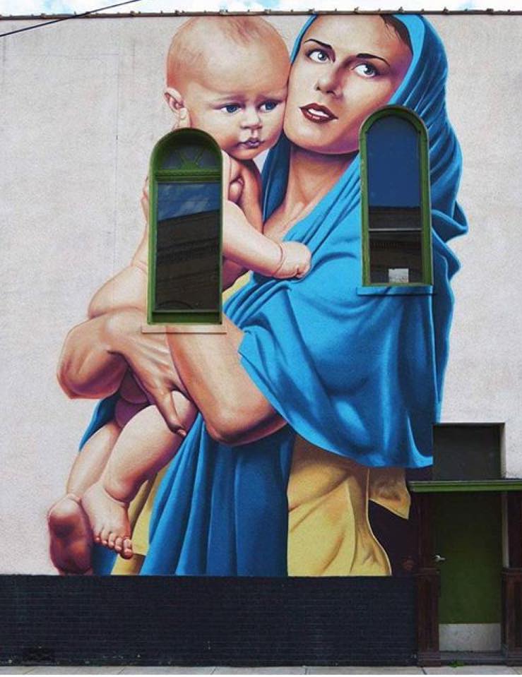 Mural of a woman embracing a baby, wrapped in a blue shawl and looking up towards the sky. Painted against the side of a historic building with a green door on the lower right hand side.